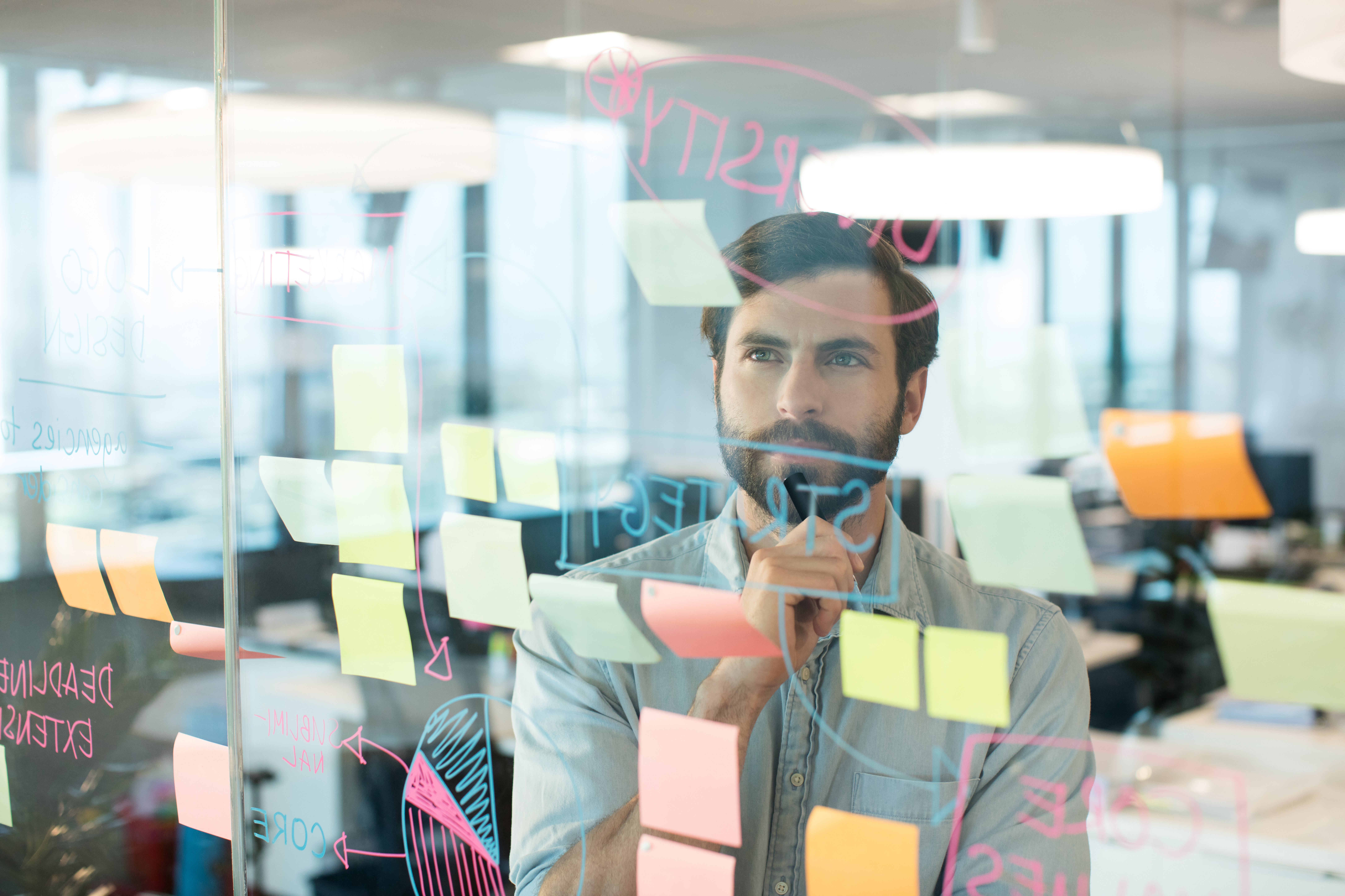 Bearded man looking through a glass planning board covered in sticky notes and diagrams in How to Hire 50+ People in a Season Without Losing Your Mind.