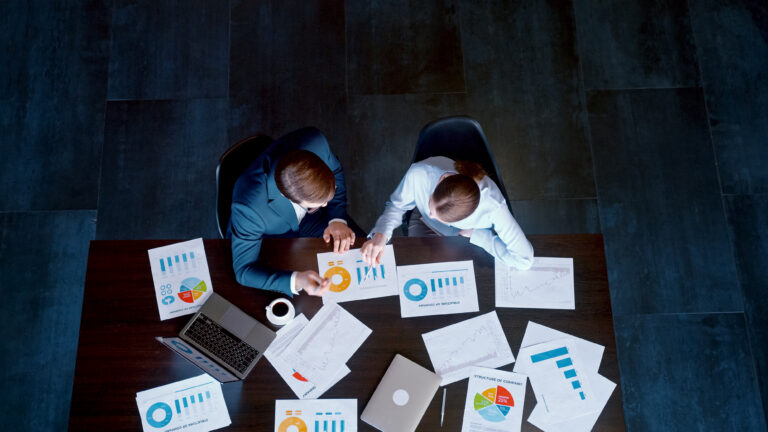 Bird's-eye view of two business professionals discussing documents, pie charts, and graphs spread across a wooden desk next to laptops